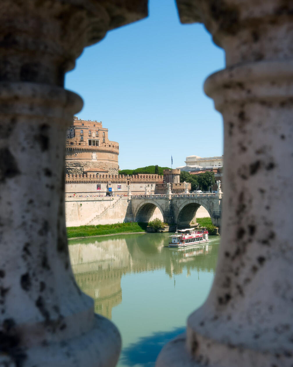 Zamek Świętego Anioła w Rzymie wraz z mostem Ponte Sant’Angelo widziane przez kamienne balustrady, na rzece płynie biały wycieczkowy statek, pod błękitnym niebem.