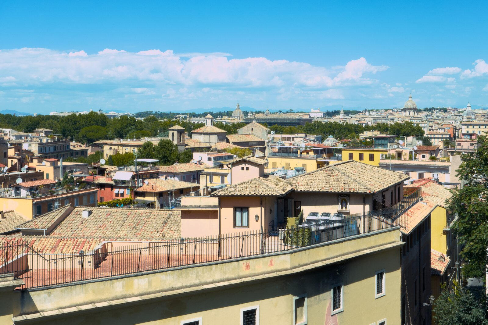 Panorama Rzymu w słoneczny dzień, dachy beżowych i żółtych kamienic z terakotową dachówką, w tle kopuły i zabytki oraz niebieskie niebo z chmurami.