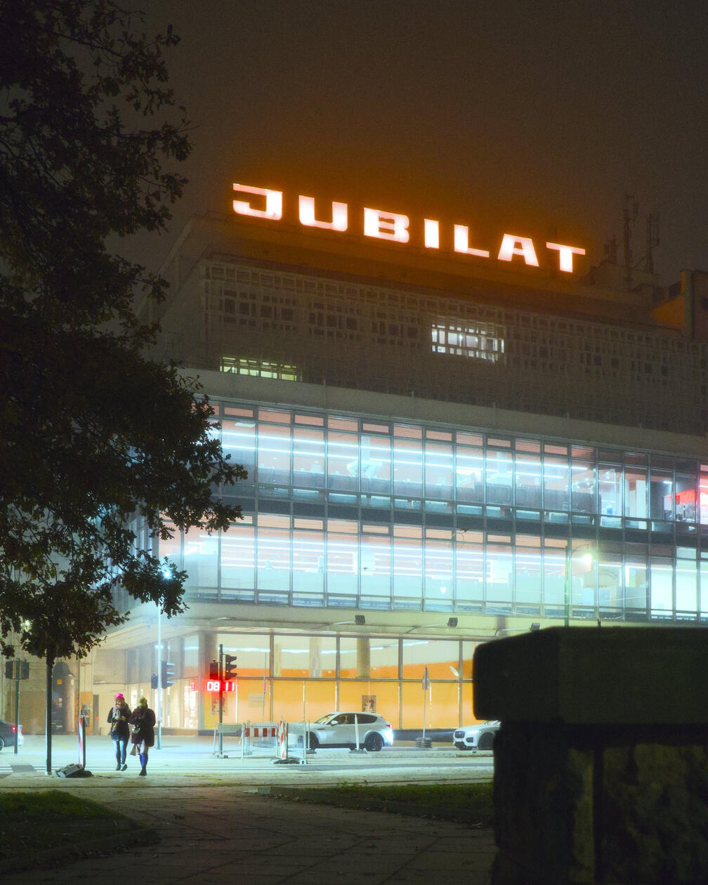 Szklany budynek Jubilat w Krakowie nocą, na dachu podświetlony na pomarańczowo neon z nazwą, po lewej stronie drzewa, w tle ulica i przechodzący ludzie.
