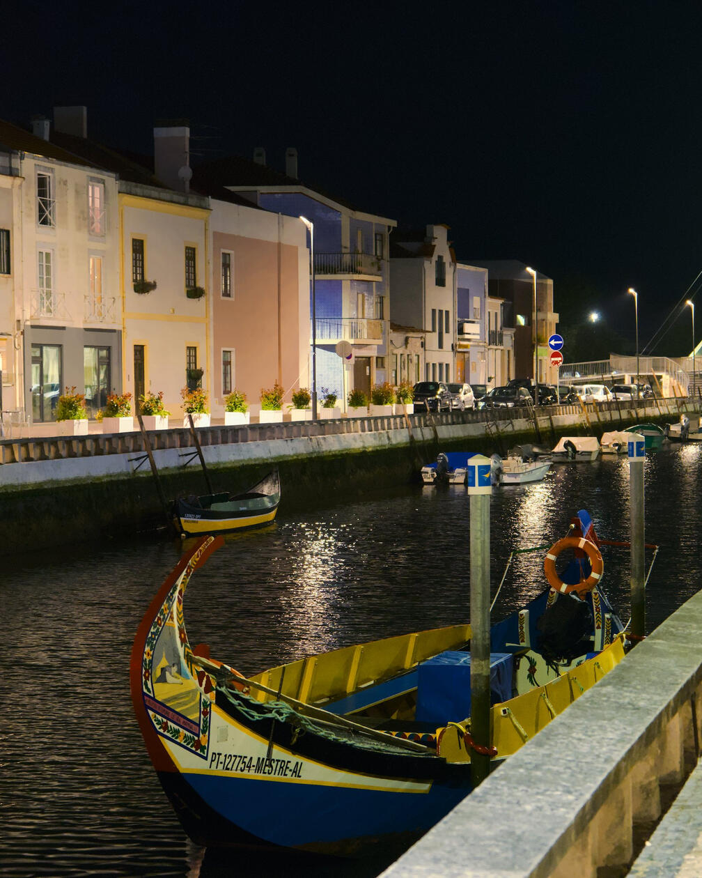 Tradycyjna kolorowa łódź cumuje przy kanale w Aveiro nocą, odbicia świateł ulicznych tańczą na wodzie, w tle rząd pastelowych kamienic.