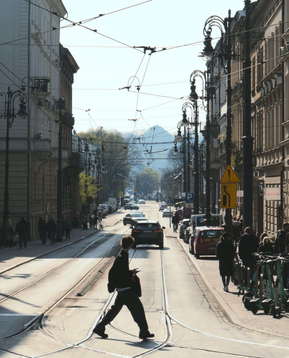 Ulica w Krakowie z zabytkowymi kamienicami, tramwajowymi torami i pieszym na przejściu, w oddali widoczny Kopiec Kościuszki w południowym świetle.