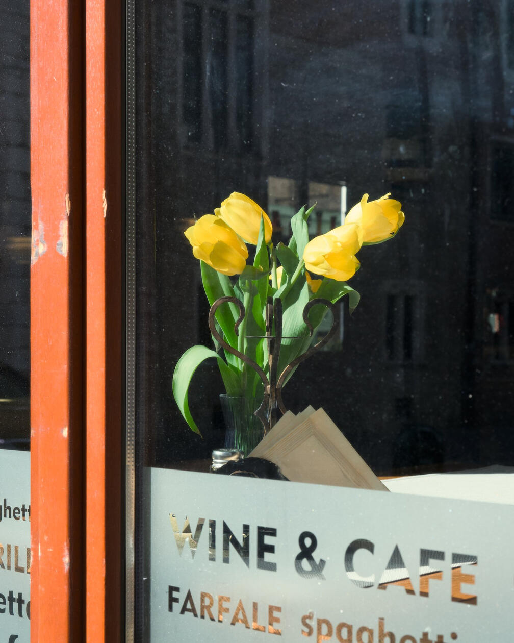Bukiet żółtych tulipanów w wazonie stoi na stoliku w oknie kawiarni, na szybie widoczne odbicia miasta i napisy 'WINE & CAFE'.