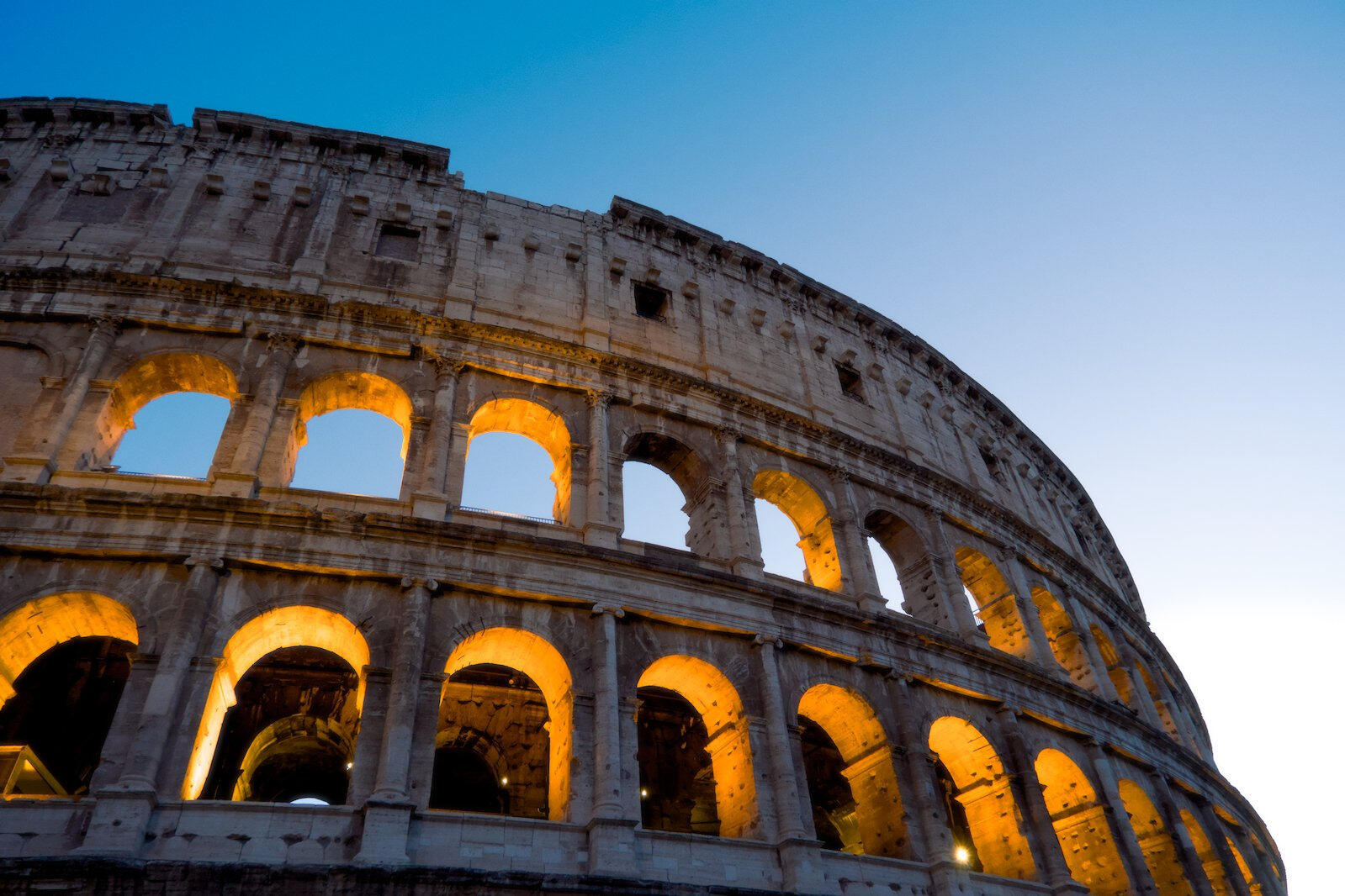 Koloseum w Rzymie o zmierzchu, z podświetlonymi na złoto łukami i niebem w odcieniu błękitu.