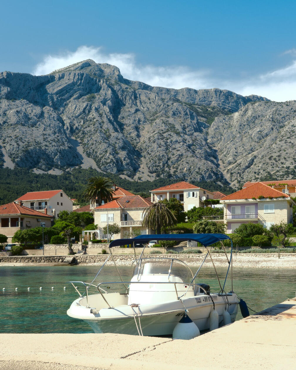 Mała motorówka zacumowana na turkusowej wodzie przy betonowym nabrzeżu, w tle śródziemnomorskie domy z czerwonymi dachami, palmy i skaliste góry pod błękitnym niebem.