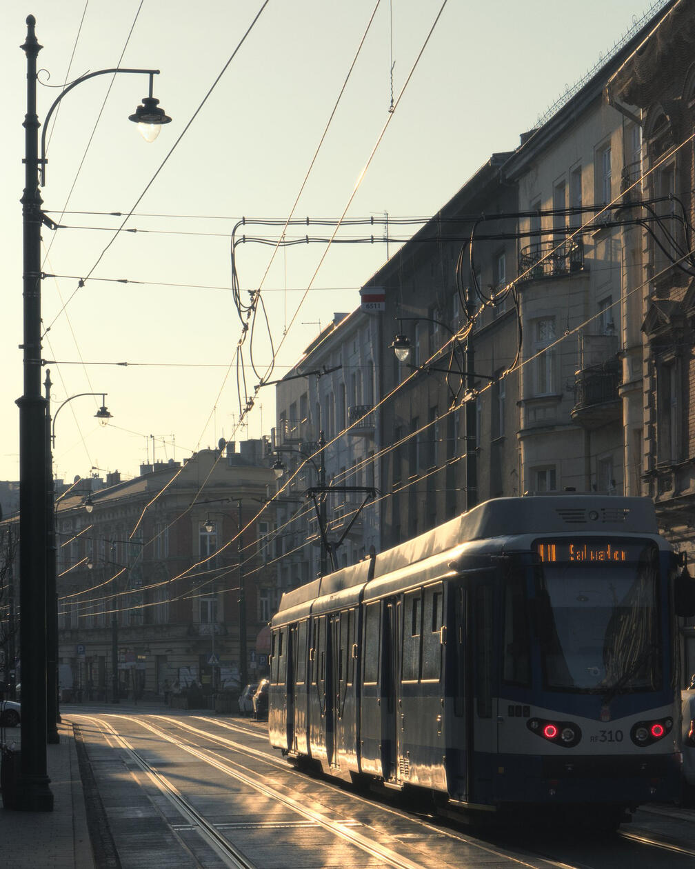 Niebiesko-biały tramwaj jedzie po jasnych torach wśród kamienic Krakowa o poranku, promienie słońca odbijają się od przewodów i okien budynków.