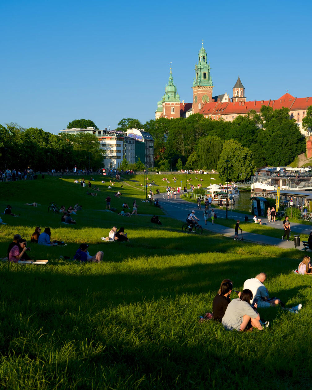 Grupy ludzi odpoczywają na zielonych wałach nad Wisłą, w tle zamek królewski na Wawelu i błękitne niebo w letnie popołudnie.