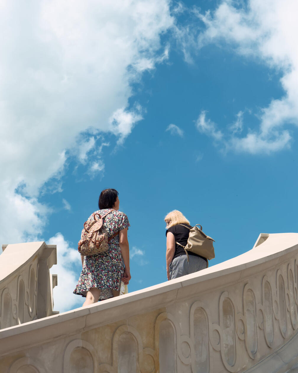 Dwie kobiety z plecakami wspinają się po ozdobnych schodach na tle błękitnego nieba i białych chmur w słoneczny dzień.