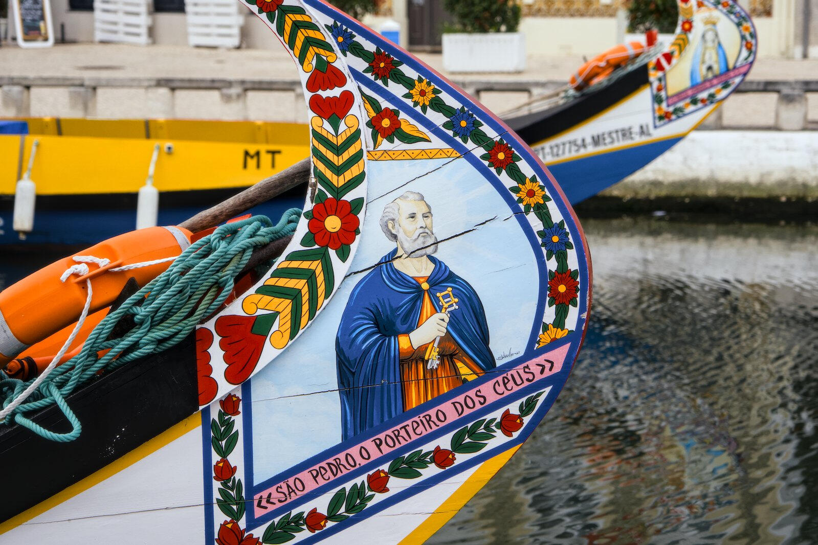 Kolorowo malowana dziób łodzi moliceiro w Aveiro z motywem świętego Piotra i napisem 'São Pedro, o porteiro dos céus', ozdobiony florystycznymi wzorami na wodzie kanału.