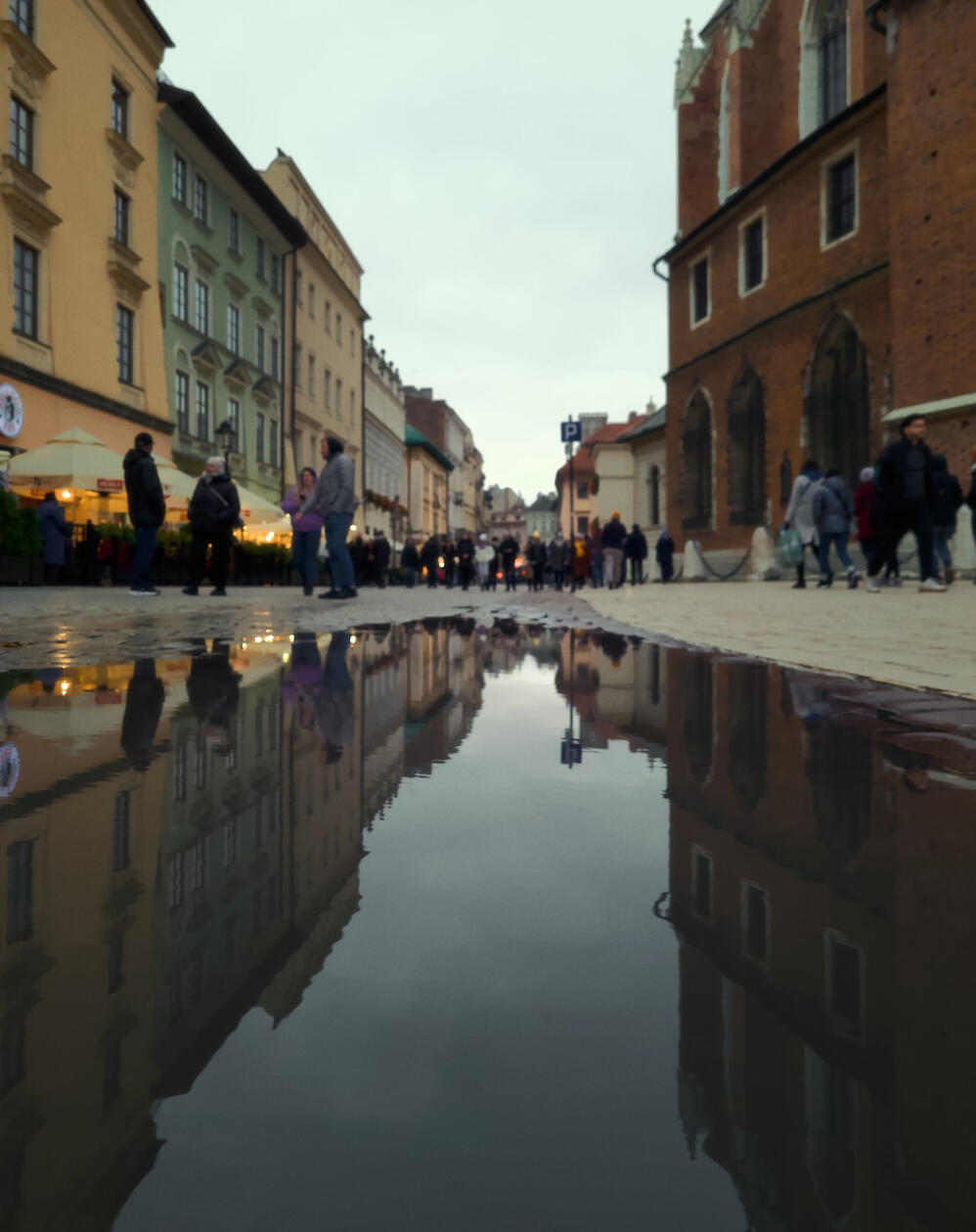 Przechodnie na krakowskiej starówce odbijają się wraz z kamienicami i kościołem w dużej kałuży na brukowanym rynku w pochmurny dzień.