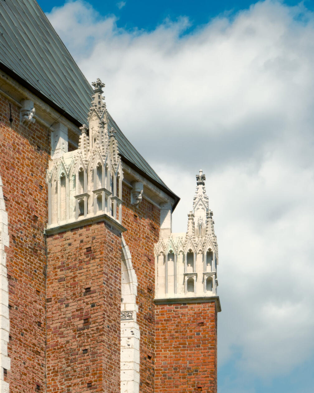 Detal gotyckich wieżyczek z białego kamienia na narożniku ceglanych murów kościoła Mariackiego w Krakowie, na tle błękitnego nieba z chmurami.