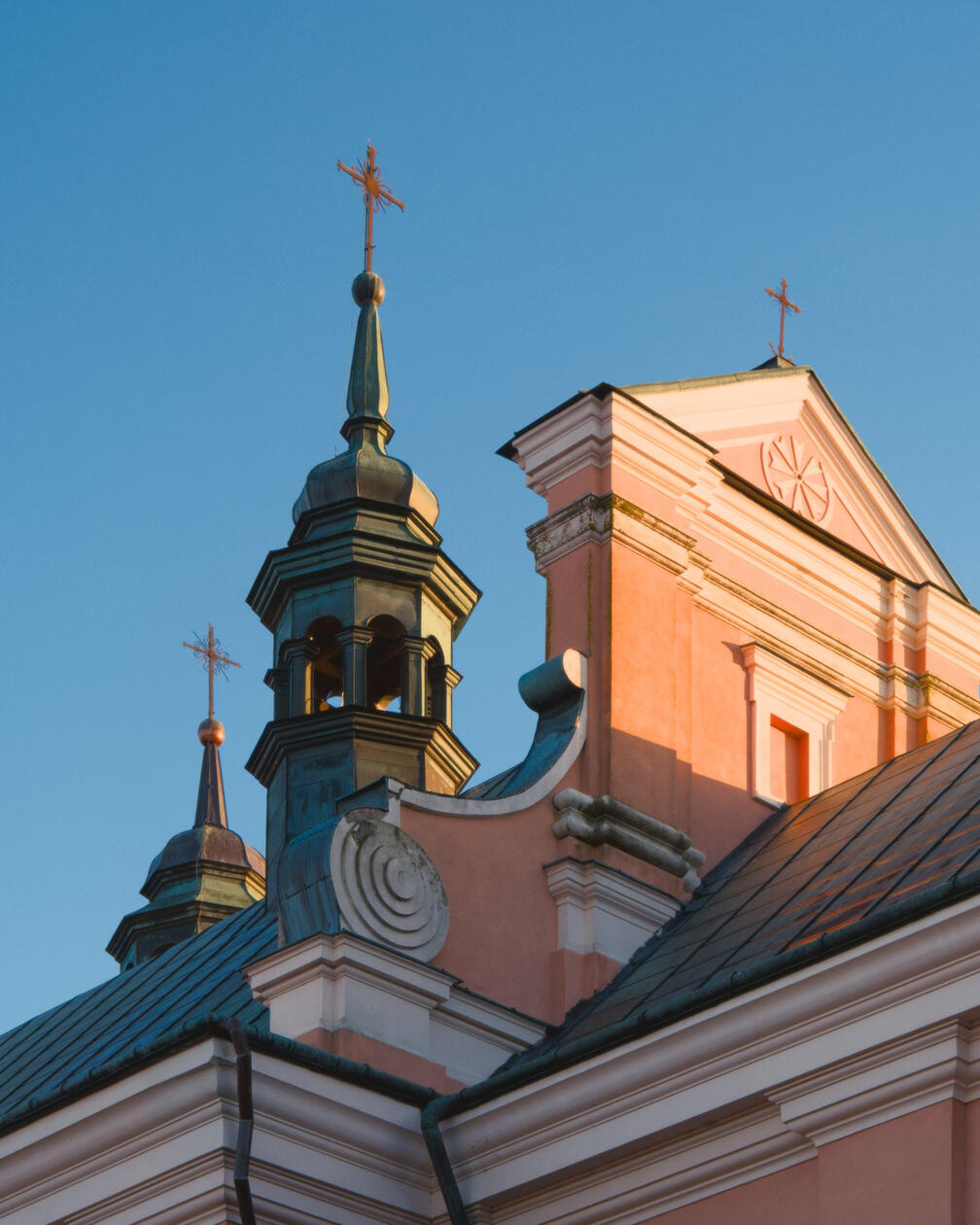 Fragment barokowego kościoła w Radecznicy widoczny w złotym świetle zachodu, z ozdobną wieżą i różową fasadą na tle błękitnego nieba.