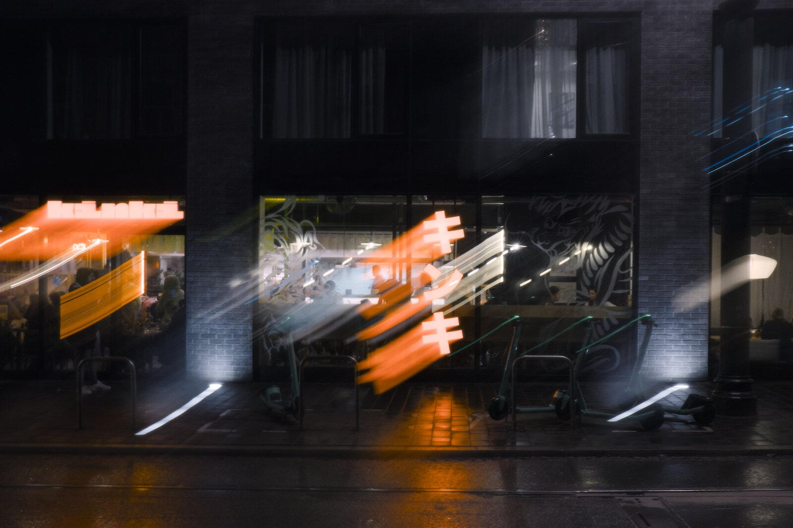 Nocna scena przed restauracją ramen z rozmazanym jasnopomarańczowym szyldem na tle ciemnej elewacji, widoczni klienci za szybą i rząd hulajnóg pod lokalem.