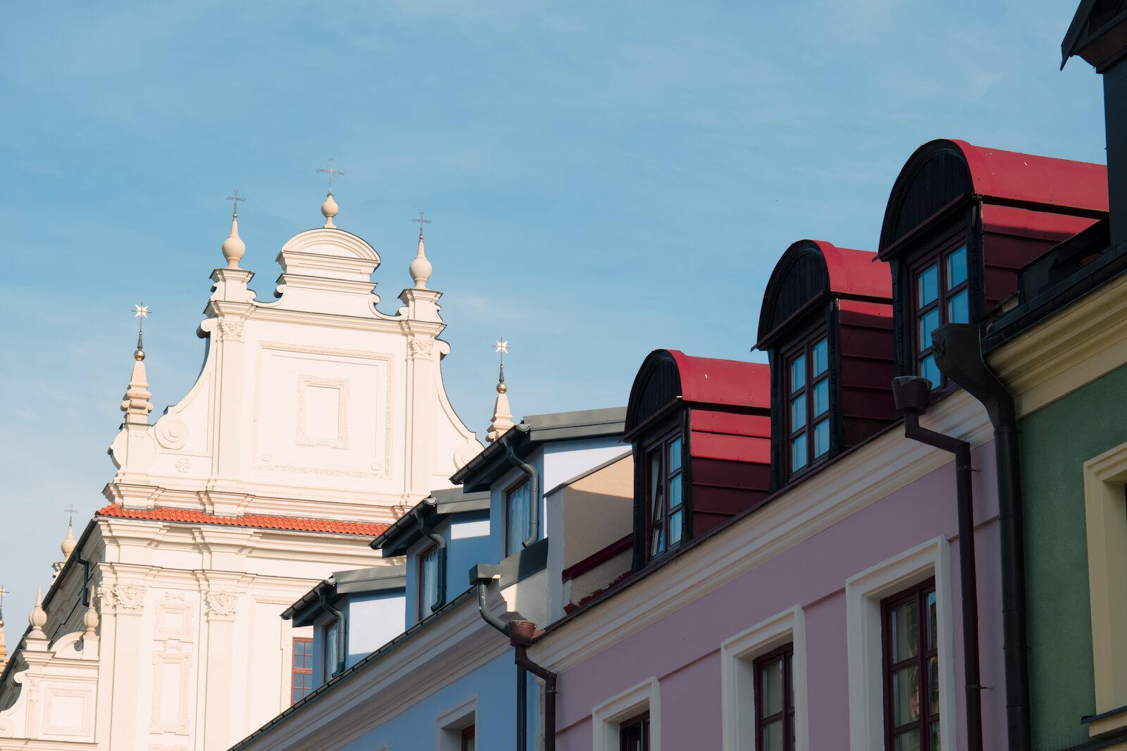 Kolorowe fasady kamienic z mansardowymi dachami i białą barokową ścianą kościoła w tle na tle jasnego nieba.
