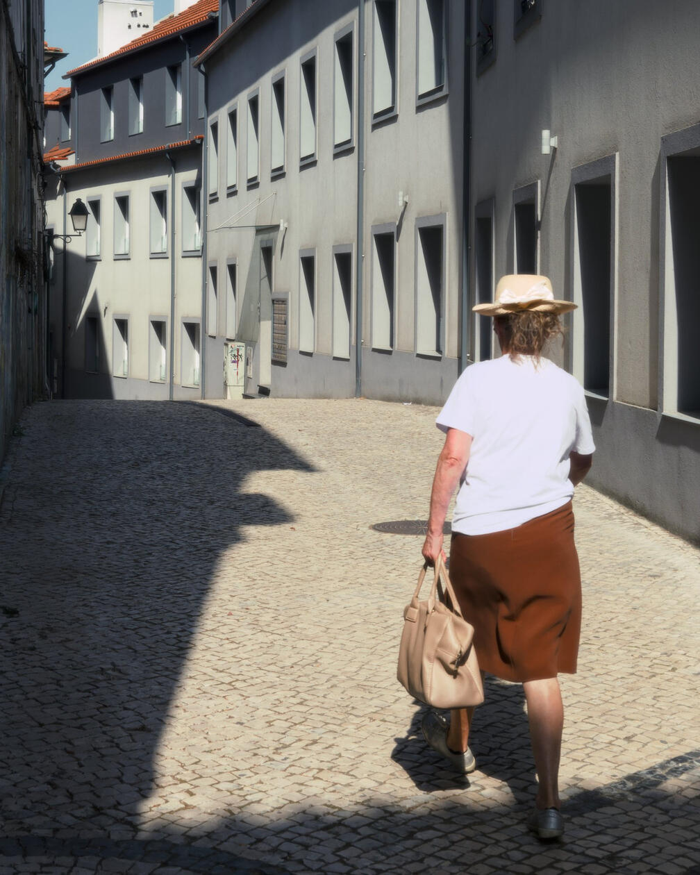 Starsza kobieta w kapeluszu z torebką idzie brukowaną uliczką wśród szarych kamienic w pełnym słońcu.