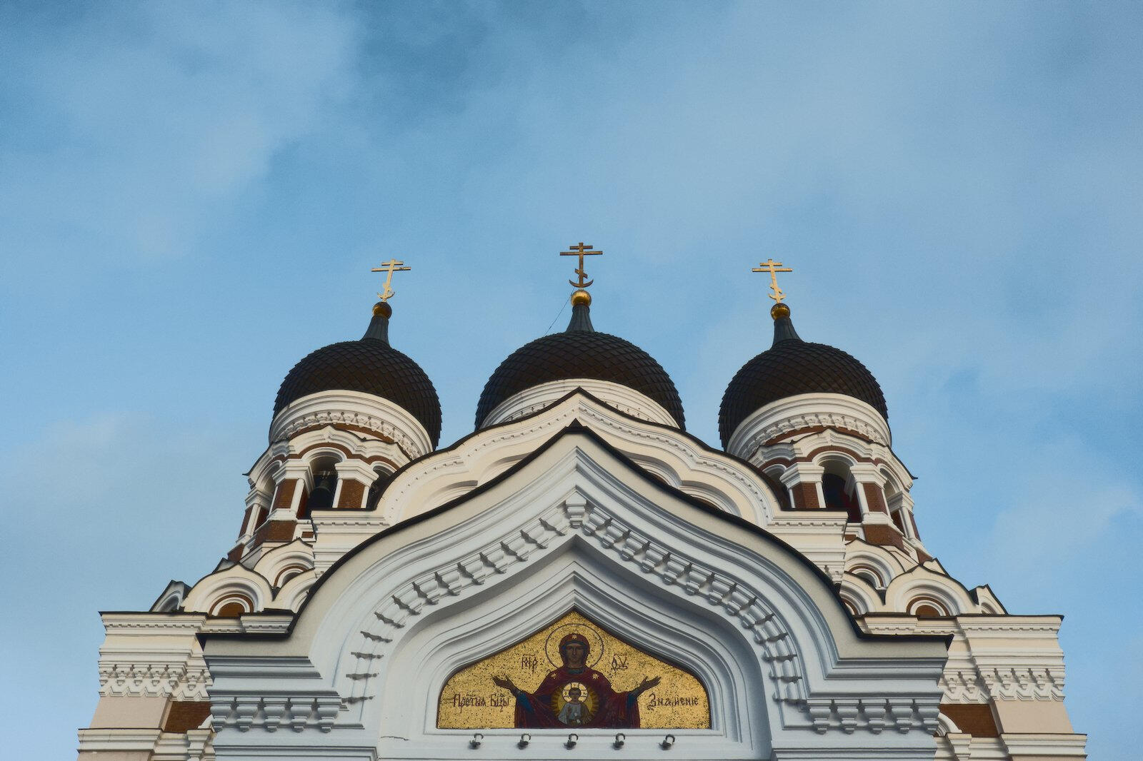 Górna część prawosławnej katedry Aleksandra Newskiego w Tallinnie, z trzema cebulastymi kopułami z krzyżami i bogato zdobioną białą fasadą na tle nieba.