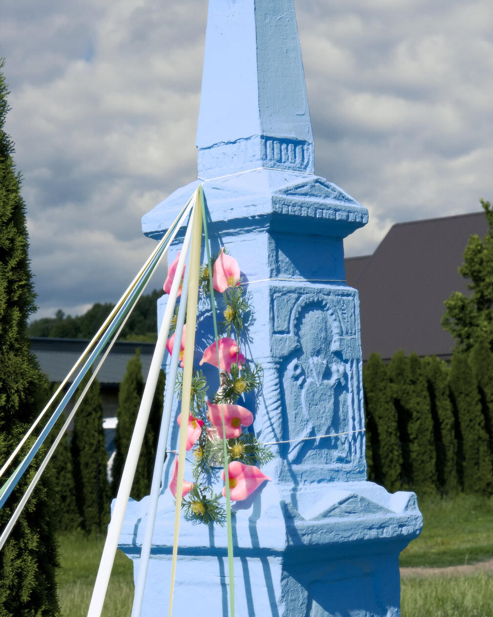 Niebieska kapliczka z płaskorzeźbą świętego, ozdobiona wiankiem z różowych sztucznych kwiatów i kolorowymi wstążkami, na tle zielonych drzew i pochmurnego nieba.