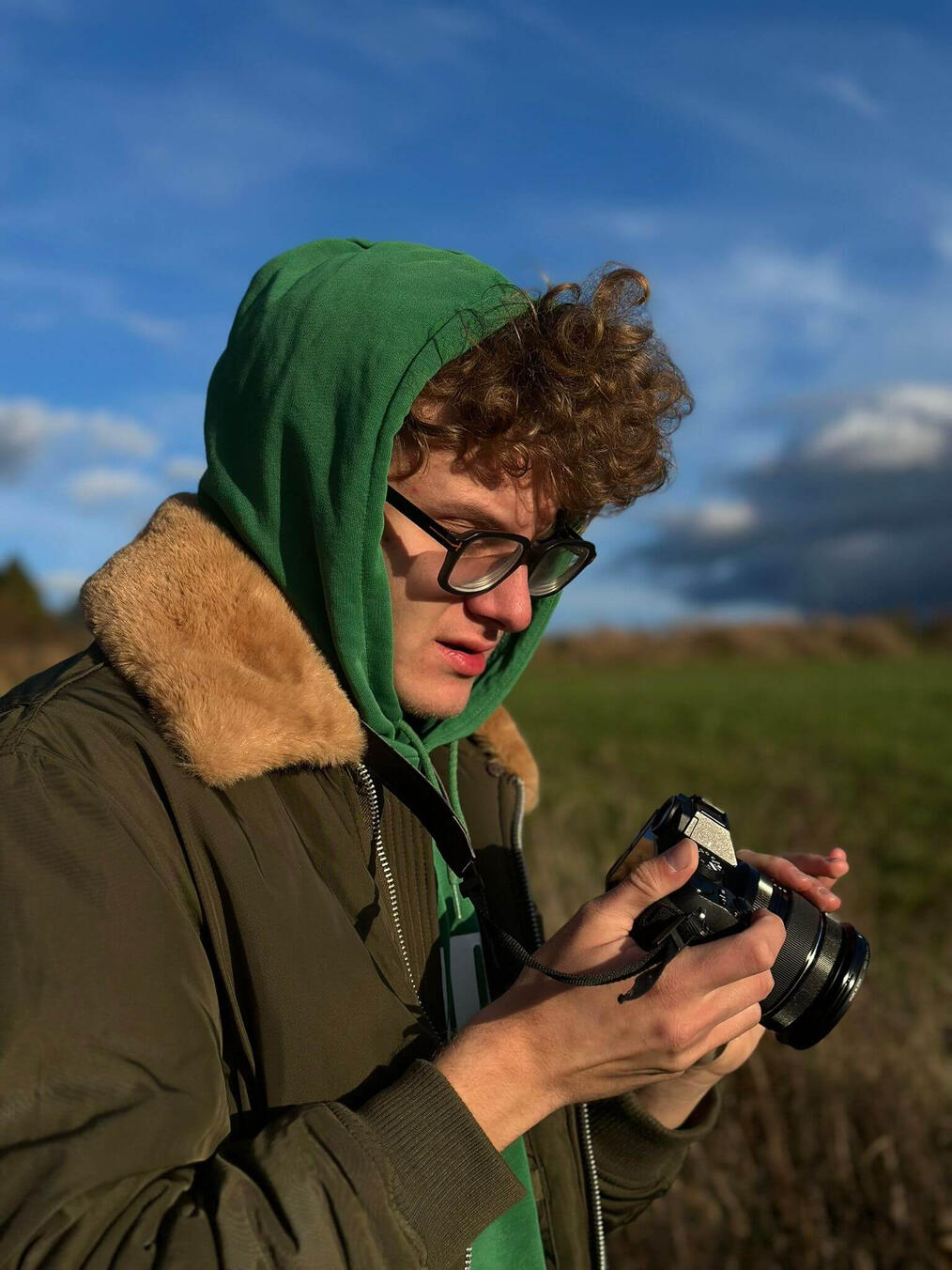 Mężczyzna z kręconymi włosami w dużych okularach, zielonej bluzie z kapturem i oliwkowej kurtce z brązowym futerkowym kołnierzykiem, trzymający i przyglądający się aparatowi fotograficznemu na tle łąki i lekko zachmurzonego nieba