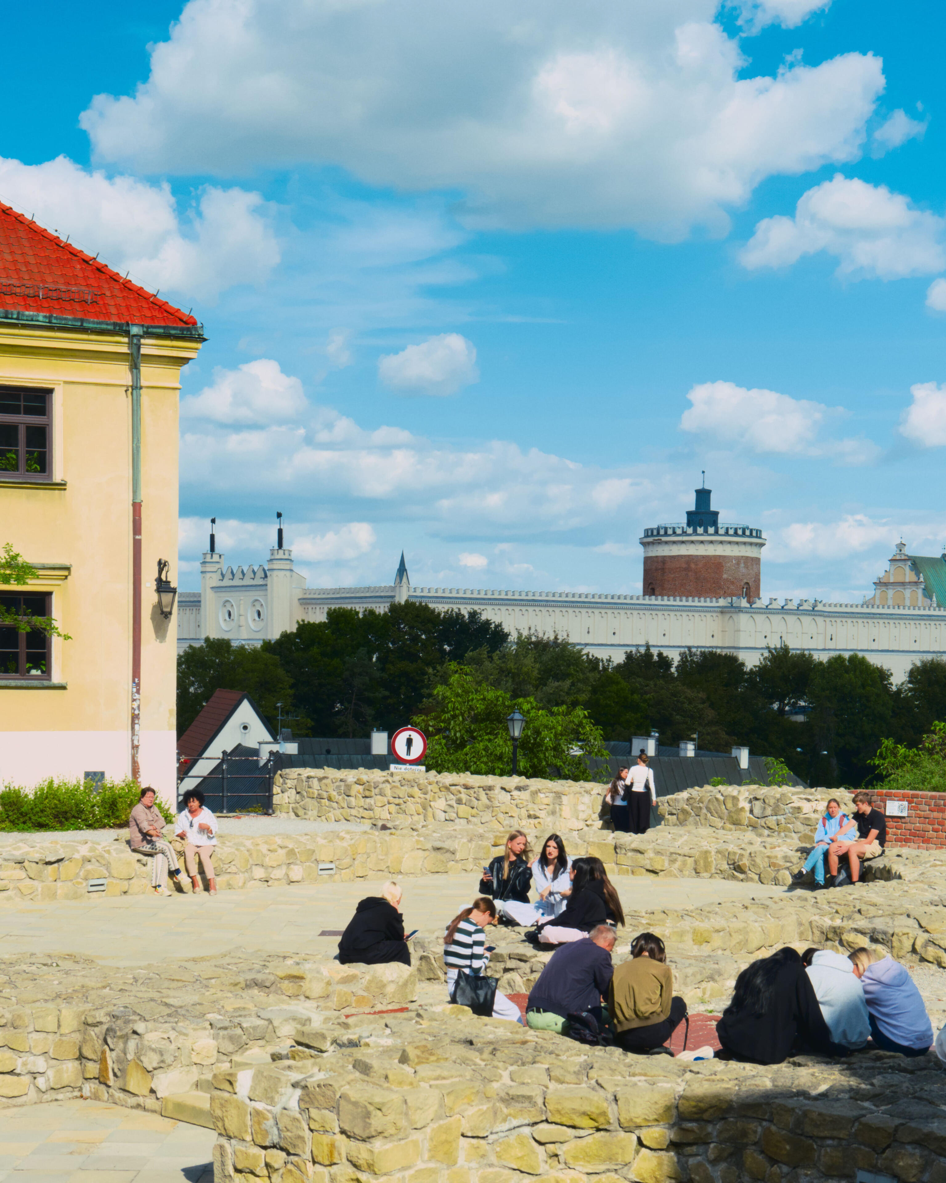 Fragment Placu po Farze w Lublinie z ruinami i murami w dolnej części kadru, na których siedzi kilka grup osób. Po lewej widoczna jest ściana i dach zabytkowego budynku. W tle widoczny jest park i Zamek Lubelski, całości dopełnia lekko zachmurzone niebo